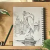 Open spiral-bound sketchbook on a wooden desk showing a pencil drawing of a large bird statue in front of a cathedral-like building, surrounded by plants, a pencil, and an eraser.