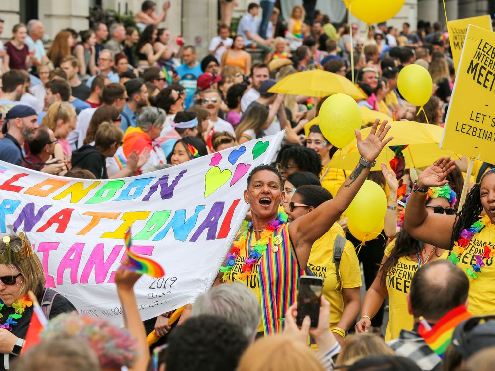 People in bright yellow shirts, including one person smiling with their arm raised, boast rainbow flags and march in support of LGBTQ people