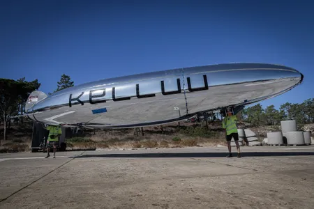 Two people standing outdoors on a paved surface holding a long, silver, cigar-shaped airship drone that features the text "KELLUU" in large black letters along its side.