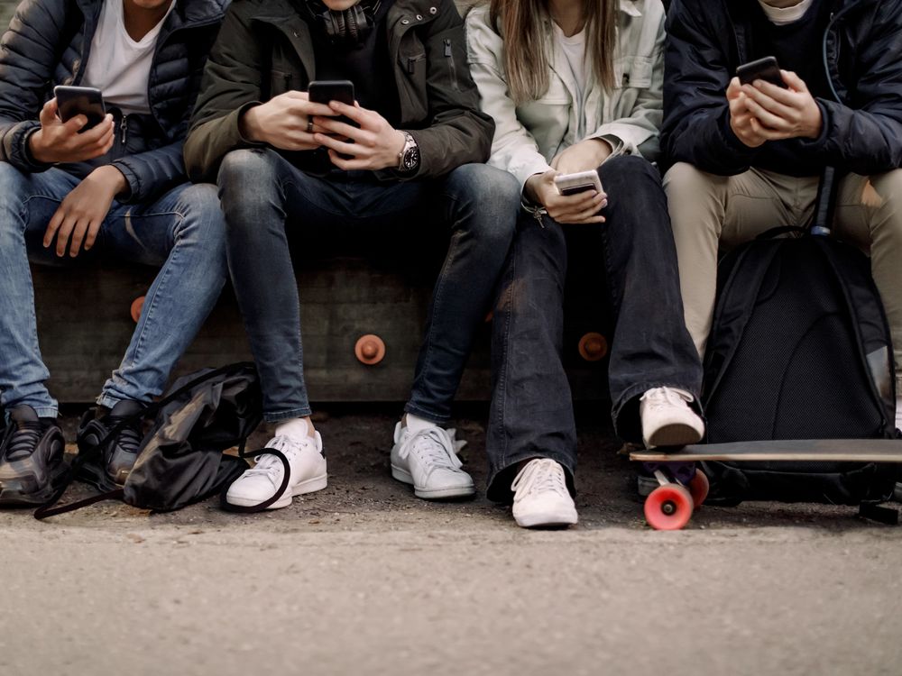 View from the waist down of kids sitting on a bench looking at their cellphones