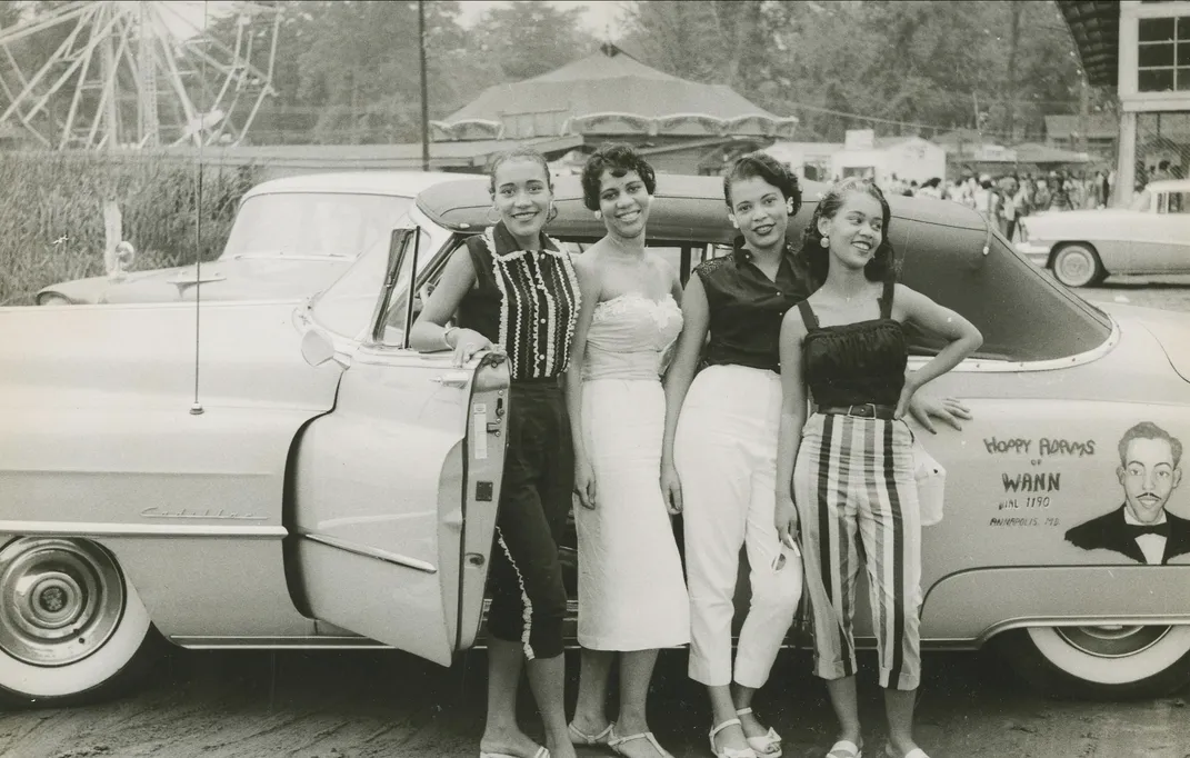 Four young women stand beside a convertible car during a visit to Carr's Beach, circa 1958