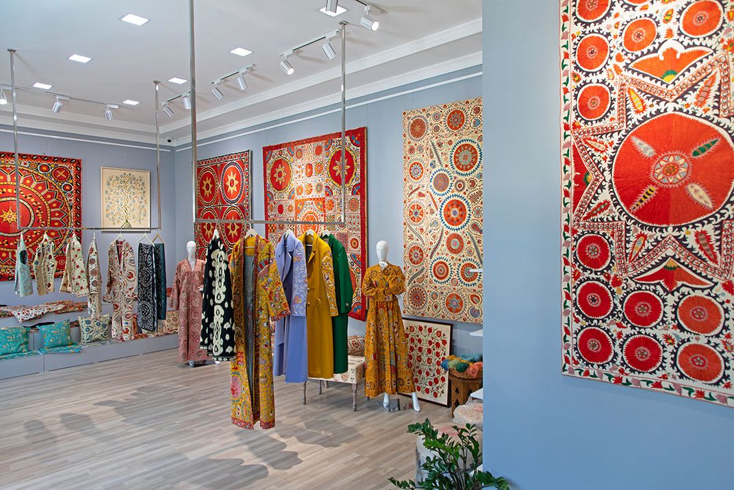 Racks of embroidered robes, dresses, and vests in a room with light blue walls decorated with red, white, and black textiles with geometric designs.