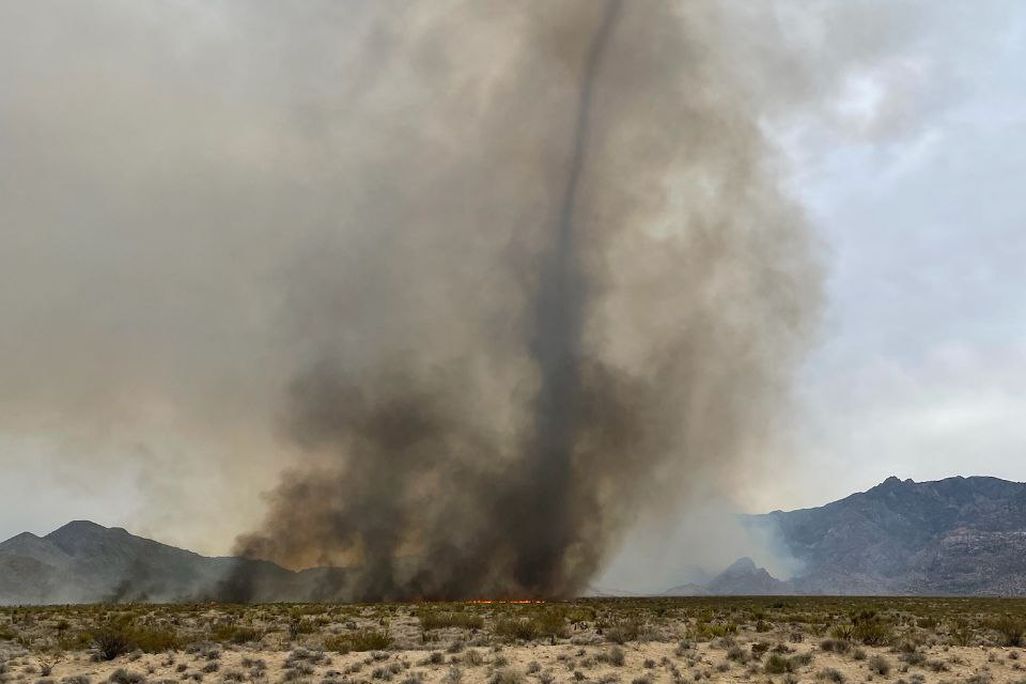 a column of black smoke rises in a desert with mountains in the background