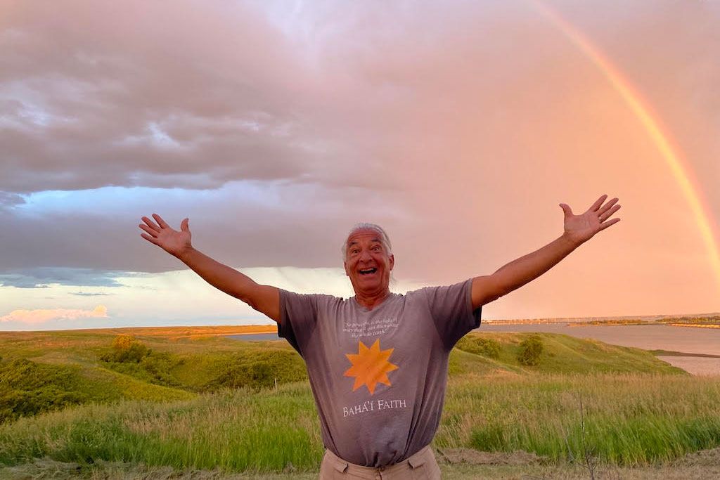 Man poses outdoors, arms outstretched, as if showing off the rainbow that stretches over his head.