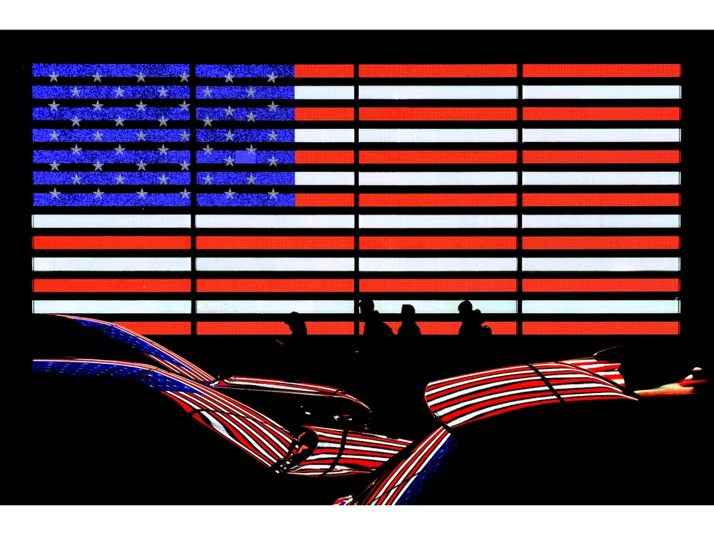 OPENER - An illuminated American flag display casts its colors onto passing cars, merging national symbolism with New York City’s constant flow of people and light.
