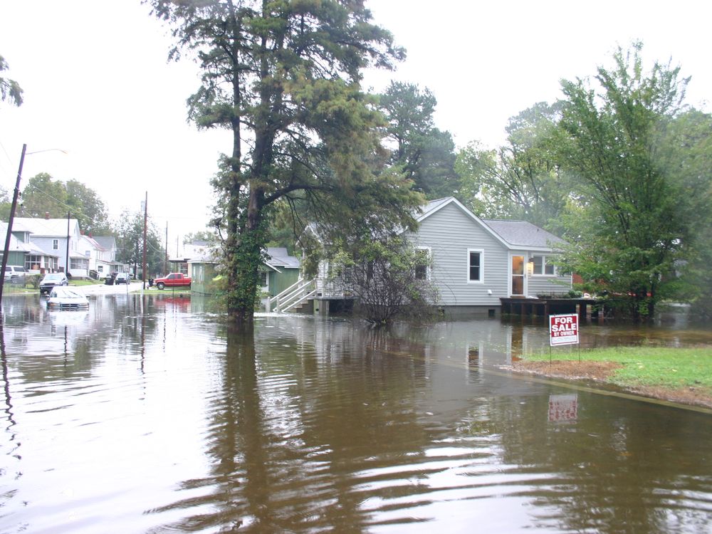 Image of a flooded neighborhood in North Carolina. The street is flooded with water and a car is halfway stuck in it. The water comes up to the houses' front porches. A "FOR SALE BY OWNER" sign is in a front yard.