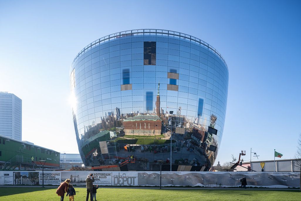 A large building shaped like a sphere with a flat top, with mirrors all around the building's surface that reflect the blue sky and white clouds
