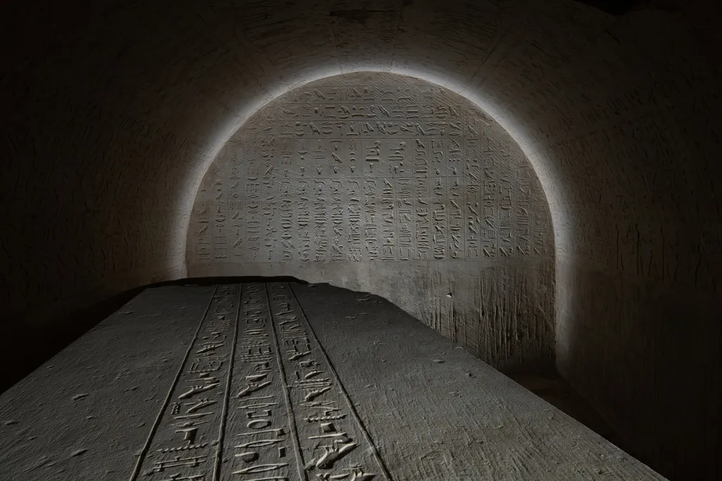 the top of a sarcophagus covered in writing with a glyph-covered wall behind it lit in an arch