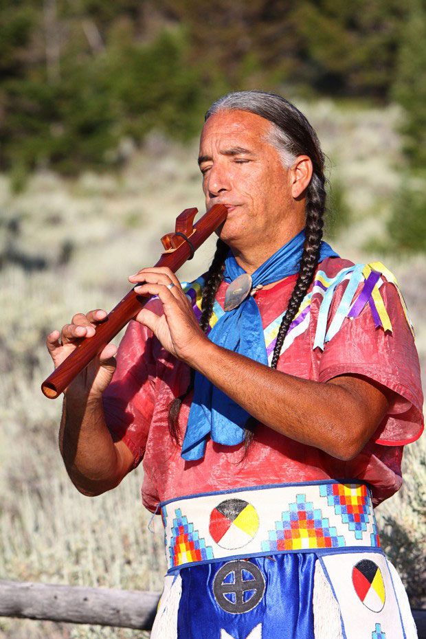Man with long braids and pink, blue, and white regalia plays a long wooden flute.