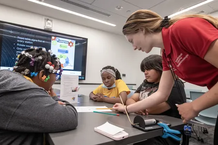 A young adult in a red polo points to a worksheet on a table. Three students are sitting at the table which has a table top rocket launcher, straw rocket, and table sign with instructions on top of it.