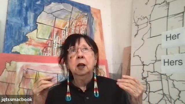 Still from a video of Jaune Quick-to-See Smith. Smith wears a black shirt with her hair pulled back, gold wire glasses, and long beaded earrings. She is sitting in front of works of art depicting maps of the United States, several in color and one in blac