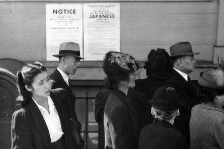 Japanese Americans stand in front of a poster with internment orders.