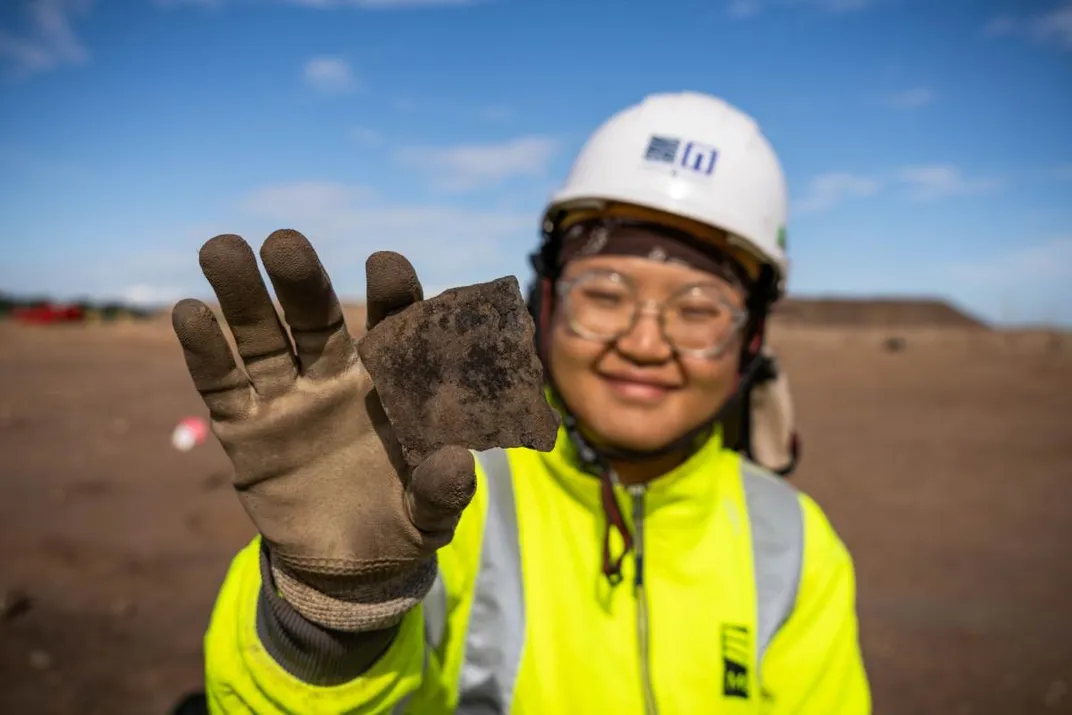 A woman in a bright yellow coat and a white hard hat holding out a piece of pottery in her hand