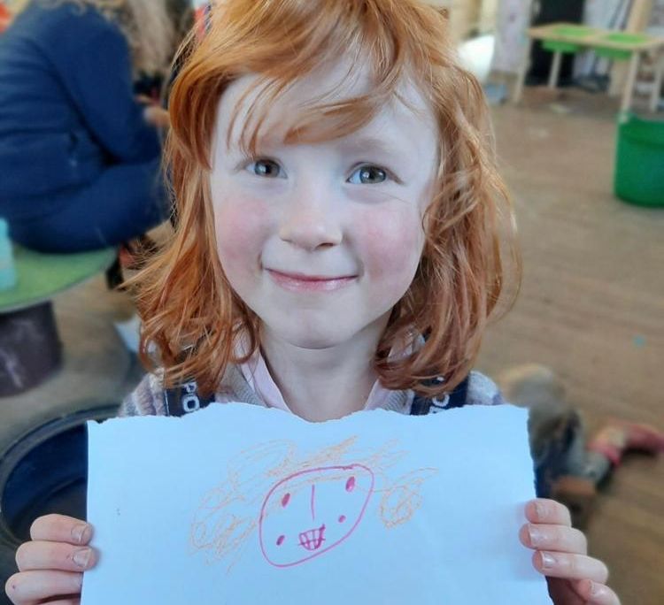 A pale, red-headed girl with bangs smiles and holds a piece of paper in front of herself, with what looks like a drawing of a person's smiling face and orange curly hair