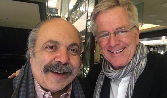 Two men stand, one wearing glasses and a scarf and the other with a mustache, smile for the camera.