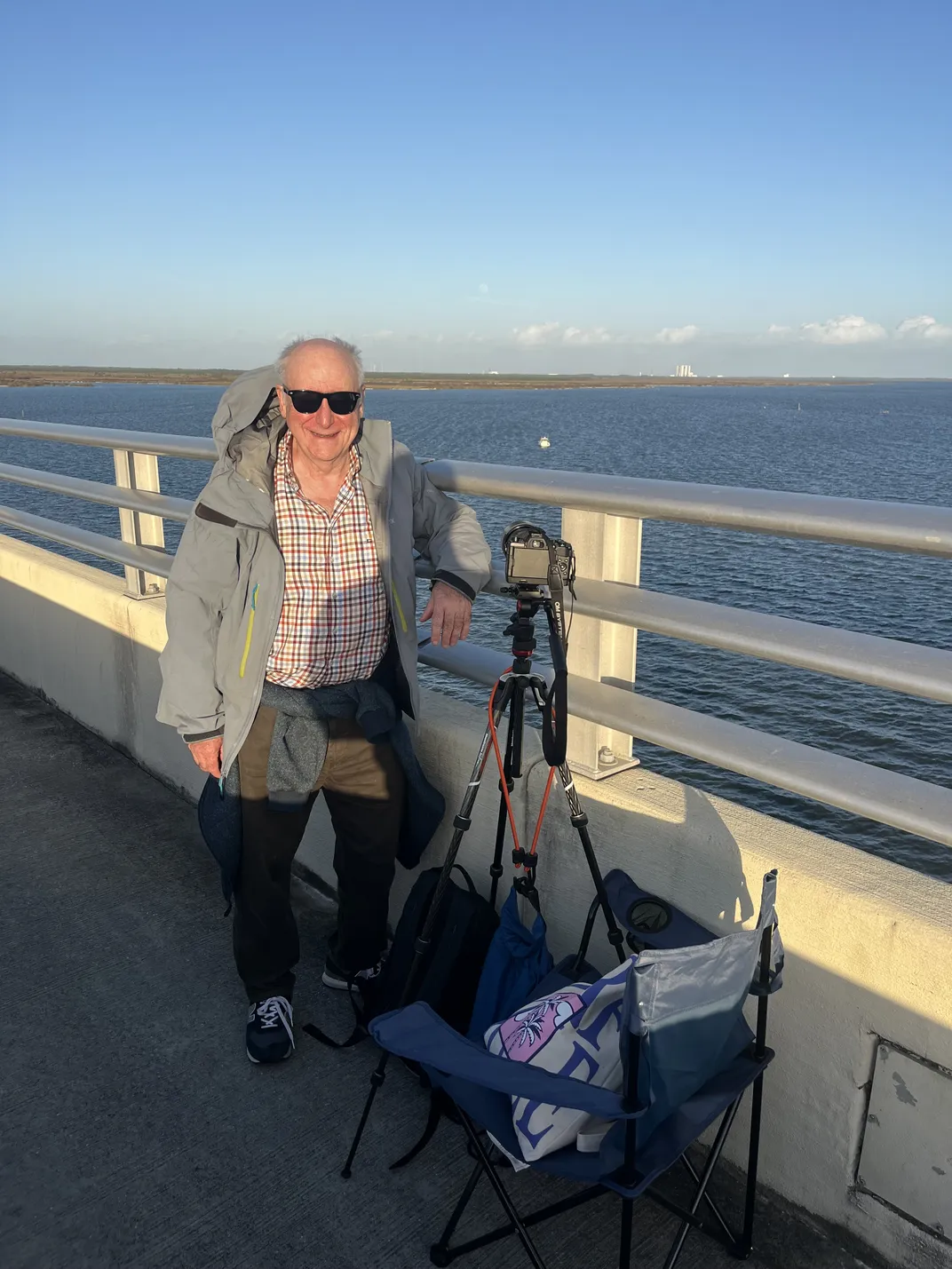 a man in sunglasses and a jacket stands near the side of a bridge with a camera and folding chair in front of him