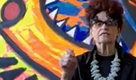 Detail of color screenshot of Judith Bernstein in front of one of her paintings wearing a black blouse and light-colored, chunky necklace holding her hand up in a fist as if making a point in conversation.