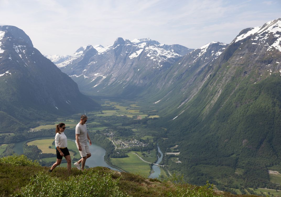 Two hikers walk along a ridge above Åndalsnes, Norway, overlooking a river valley, green farmland, and rugged snow-capped mountains.
