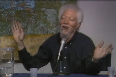 Benny Andrews sits at a table with a microphone, cups, and a pitcher of water with a yellow, green, and blue abstract painting hanging behind him. He is wearing a black button down shirt and raising his hands while talking.