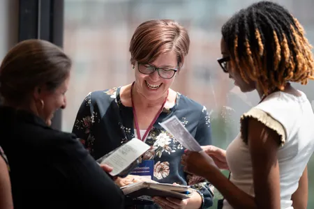 Three women stand in a circle laughing and discussing printed handouts