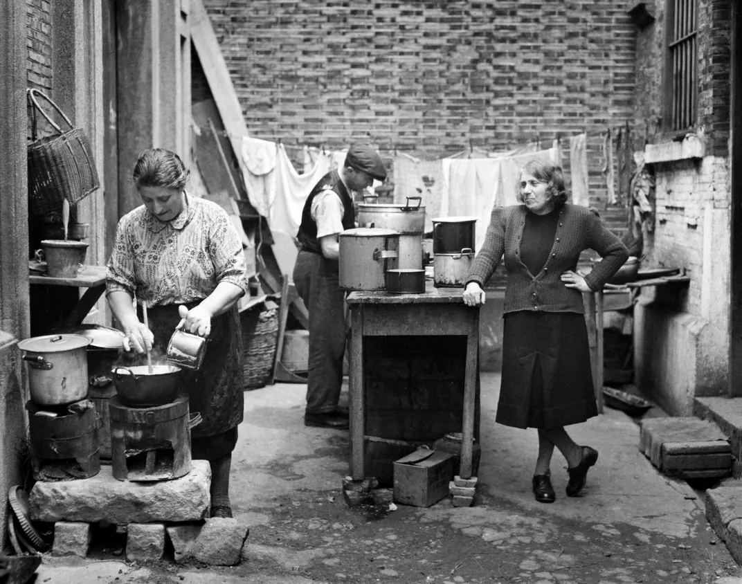 A black and white image of three people working in an outdoor kitchen, cooking with laundry hanging behind them