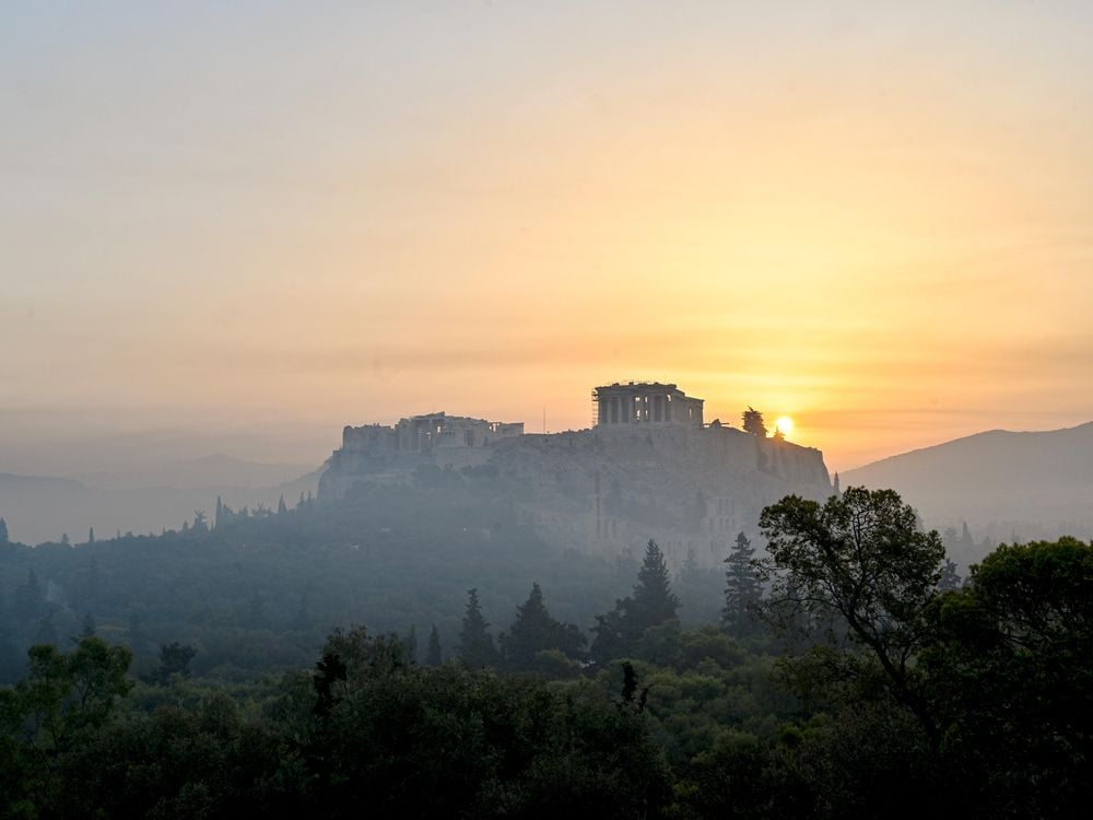 A view of the Parthenon in the distance on a hilltop, with the sun low in the sky behind a haze of smoke