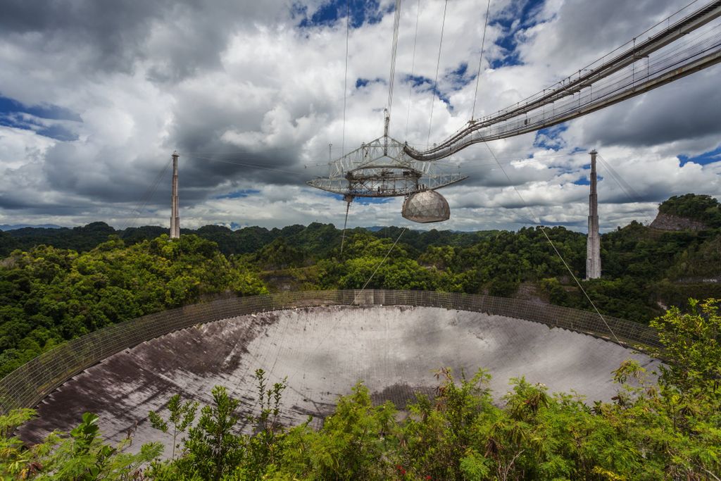 A large metal saucer-shaped object, with a dome attached to its right side, is suspended high above a large dome underneath, the antenna. Behind, an expansive view of blue sky with white clouds and rainforest gives a sense of the telescope's huge size