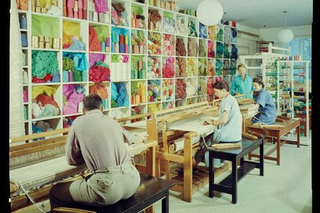 A smiling Dorothy Liebes stands in her studio in her blue apron, supervising two young women sitting at looms with spools of colorful thread and skeins of yarn close at hand.