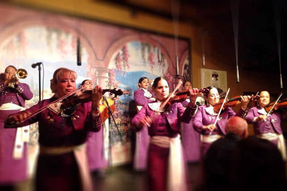 An all women Mariachi band, outfitted in matching purple and white traditional clothing, performs together in front of a painted mural depicting an archway facing the ocean.