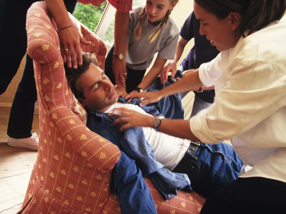 a man fainted in an armchair with four people reaching out to help him