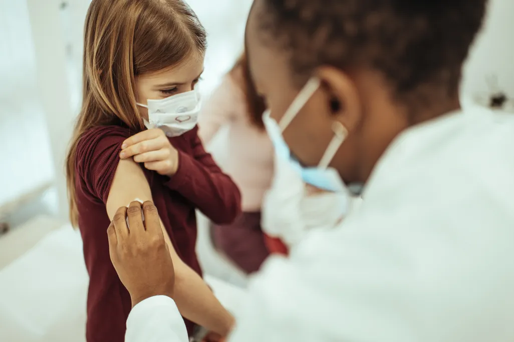 A female doctor wearing a mask vaccines a young girl wearing a mask