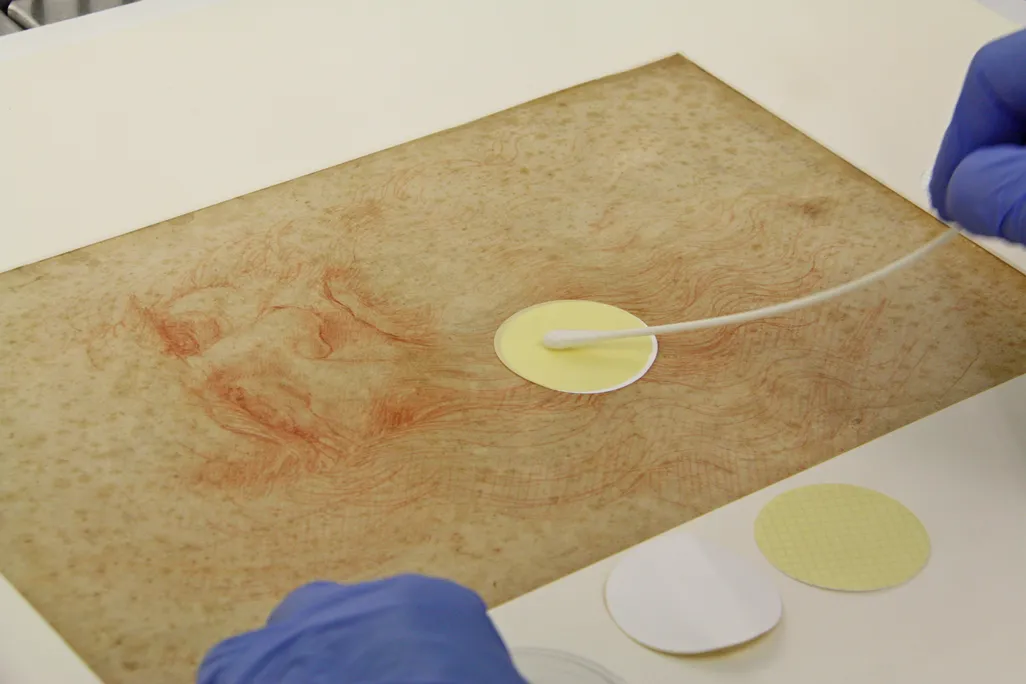 A pair of blue gloved hands gently wipe a bendy q-tip apparatus over a small circle of yellow paper that appears to be resting on the surface of the sketch, which is about the size of a normal sheet of paper