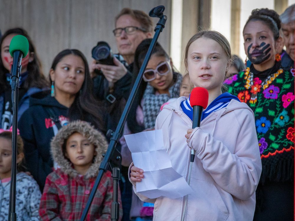 young blond girl in white coat with blue color holding a red microphone with speech in hand speaking in front of young kids