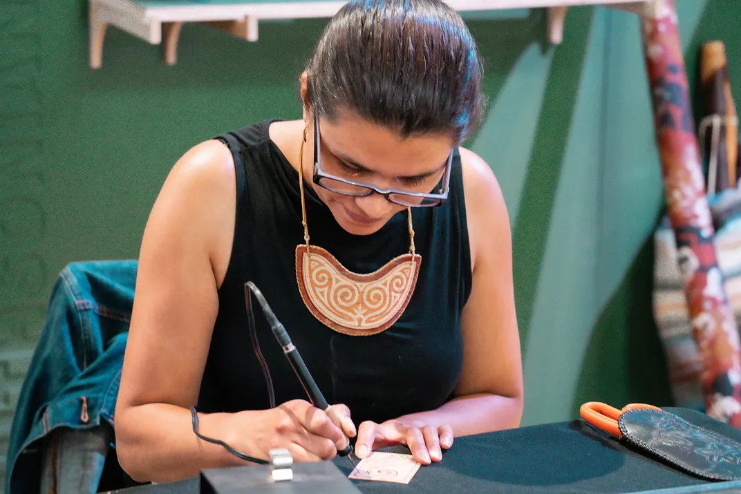 A woman wearing glasses and black tank top focuses on her work with a pen-like wood-burning tool.
