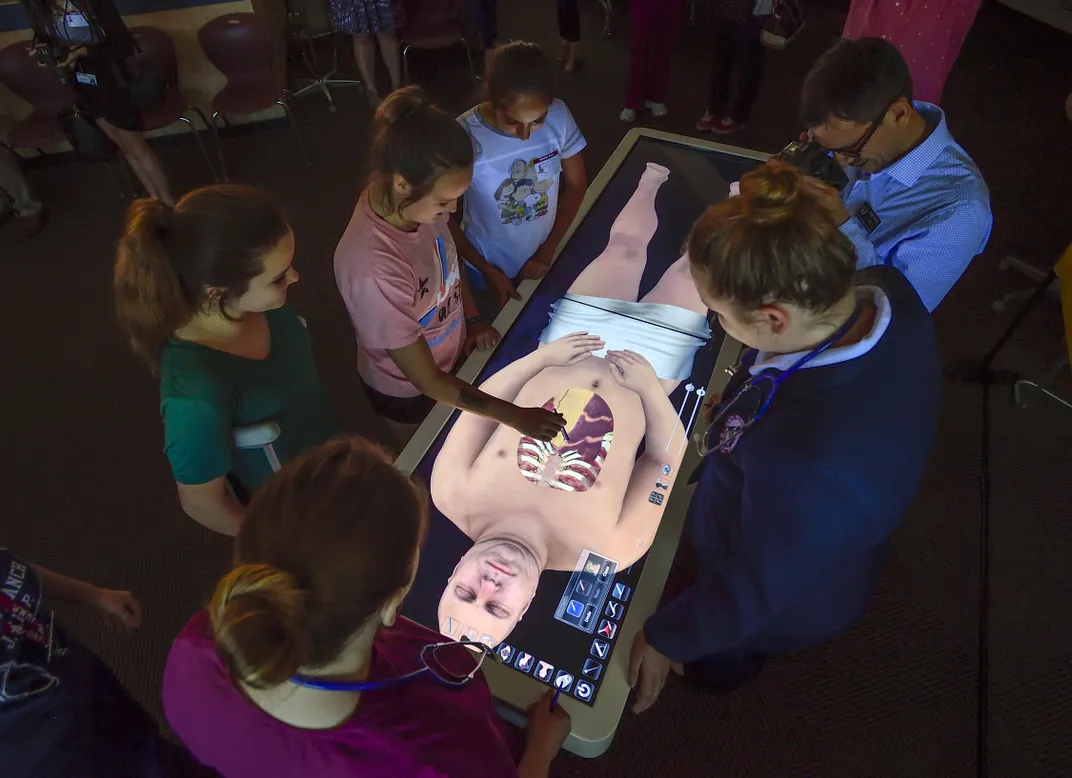 young students stand around a large horizontal screen that shows a dead man, some virtual organs visible through his ches