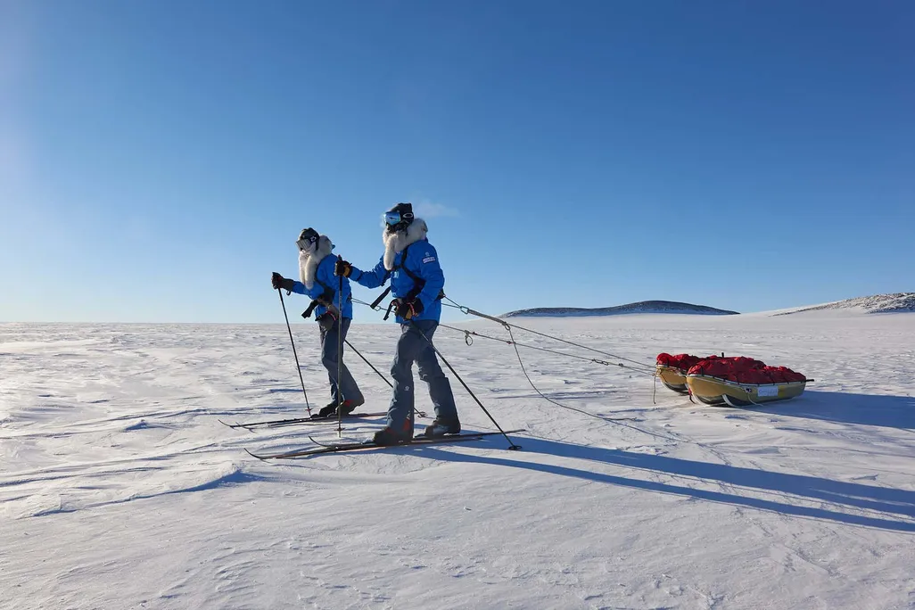 Two men in winter clothes and skis pull sleds across and frozen landscape