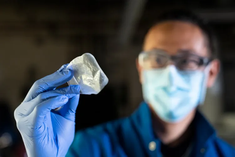 A man wearing a mask and glasses and blue gloves holds up a piece of plastic film