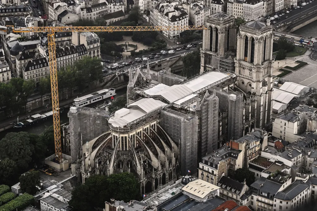 An aerial view of the exterior of the cathedral, its damaged roof mostly covered by white tarp. An enormous crane looms over the building