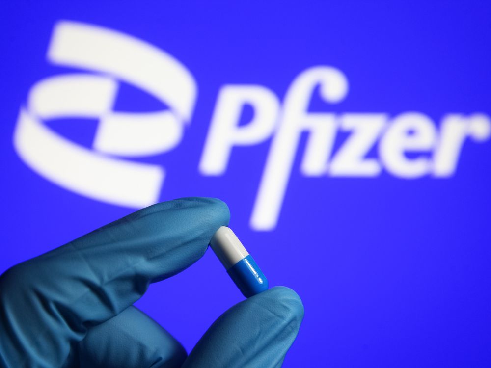 A gloved hand holds a pill in front of the Pfizer logo