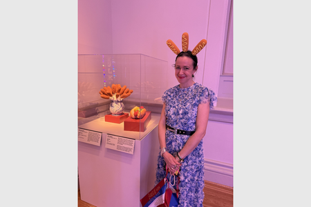 Sarah Rogers Morris wearing a headband with three corndogs poking out stands behind an exhibition case with a vase containing corndogs and a ceramic hot dog.