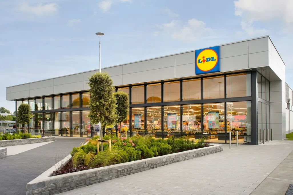 An image of a brightly lit new Lidl store; a large square building with the store's logo, a blue square with a yellow circle and blue and red block text that reads LIDL; surrounded by pavement and a parking lot