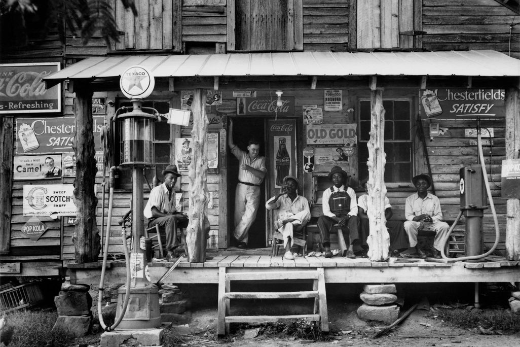 A group of people sit on the porch of a general store, surrounded by signs for products like Coca Cola and Chesterfield's cigarretes; most look at the camera 