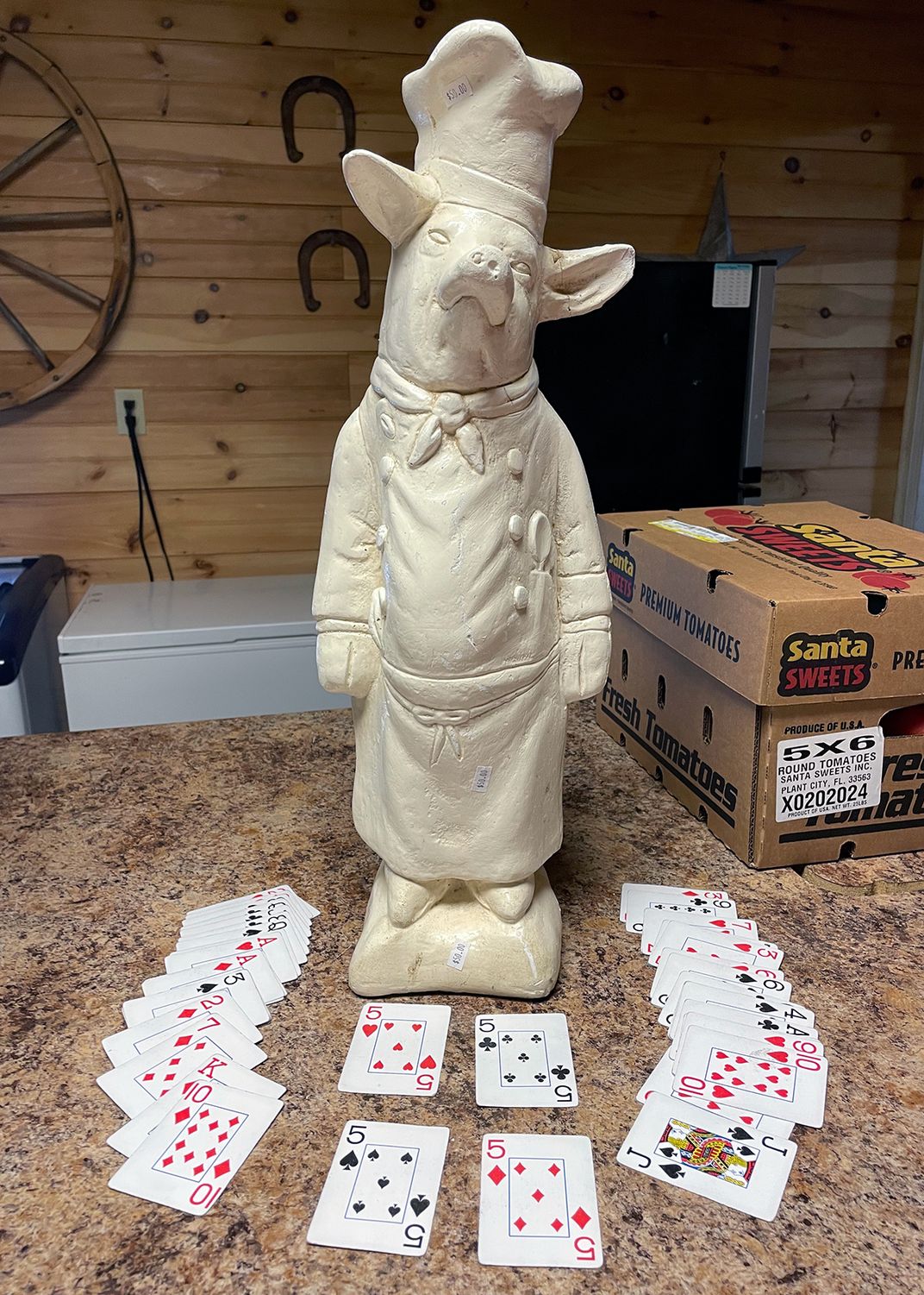 A white figurine of a pig standing on its hind legs, wearing a chef's hat and coat. On the table around it, a deck of playing cards are arranged, with fives of all suits in front.