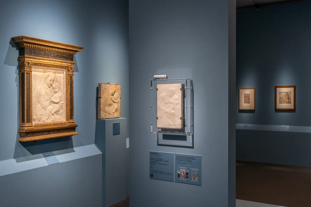 Three works hang against a blue wall, all carved reliefs of a mother and child