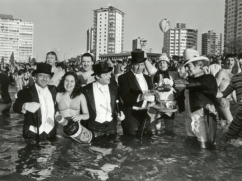 people, some in formal wear others in costumes or swimsuits, pose for a photo in water in front of a city skyline