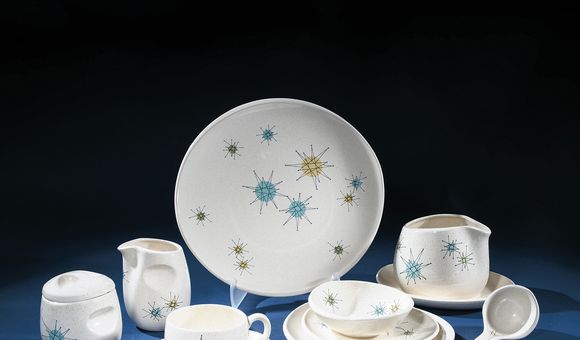 Photographed against a dark blue background is a studio image of a set of cream-colored ceramic dishware bearing a pattern of star-shapes in blue, yellow, and green. There are several pieces, including a platter, a gravy boat with a ladle, a creamer and s