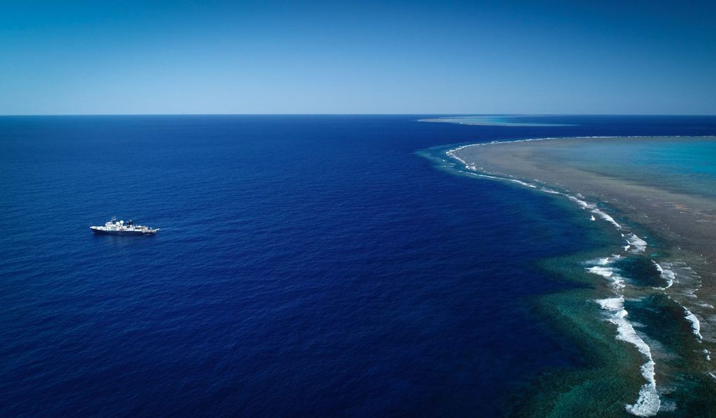 A photo taken from high above (likely by drone) shows the vast, bright blue ocean. The research vessel is on the left side. A long green and beige stretch of shallow coral reefs is on the right side. The horizon, where the deep blue ocean meets the lighte