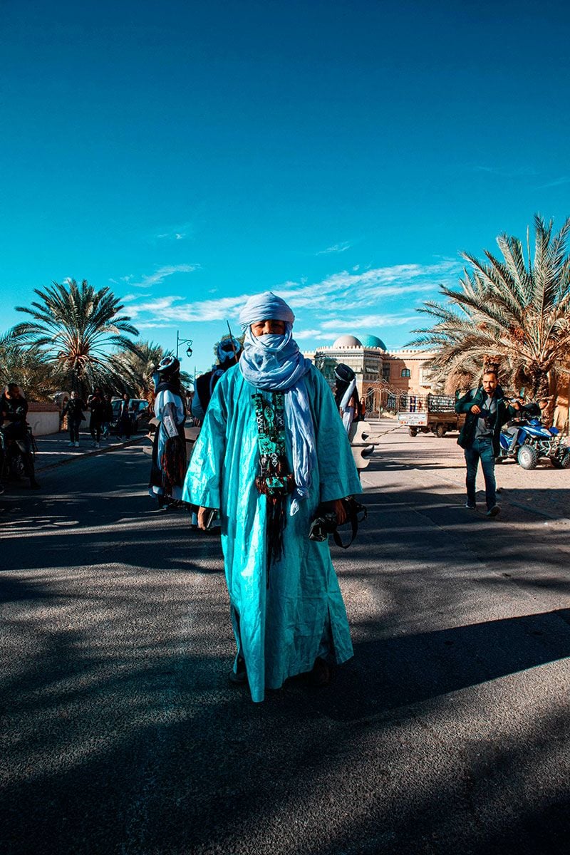 A man stands in a palm-tree lined street wearing a turquoise tunic and light blue headscarf.