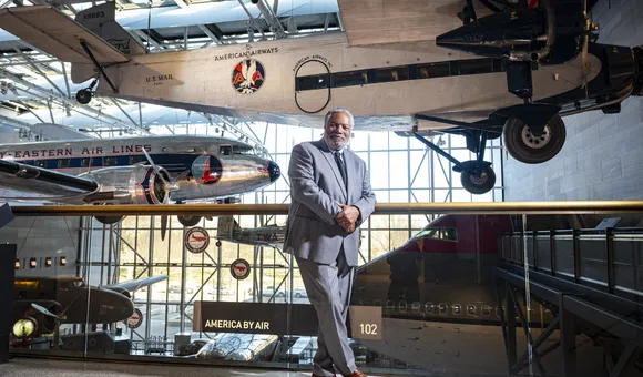 A Black man in his early 70s, wearing a blue suit and brown dress shoes, stands on a mezzanine overlooking a museum gallery displaying at least five airplanes.
