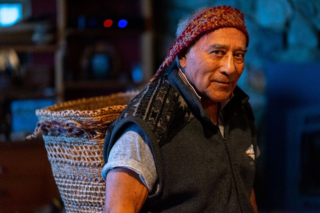 A man poses, wearing a fleece vest and a basket on his back, balanced by a woven strap across his forehead.